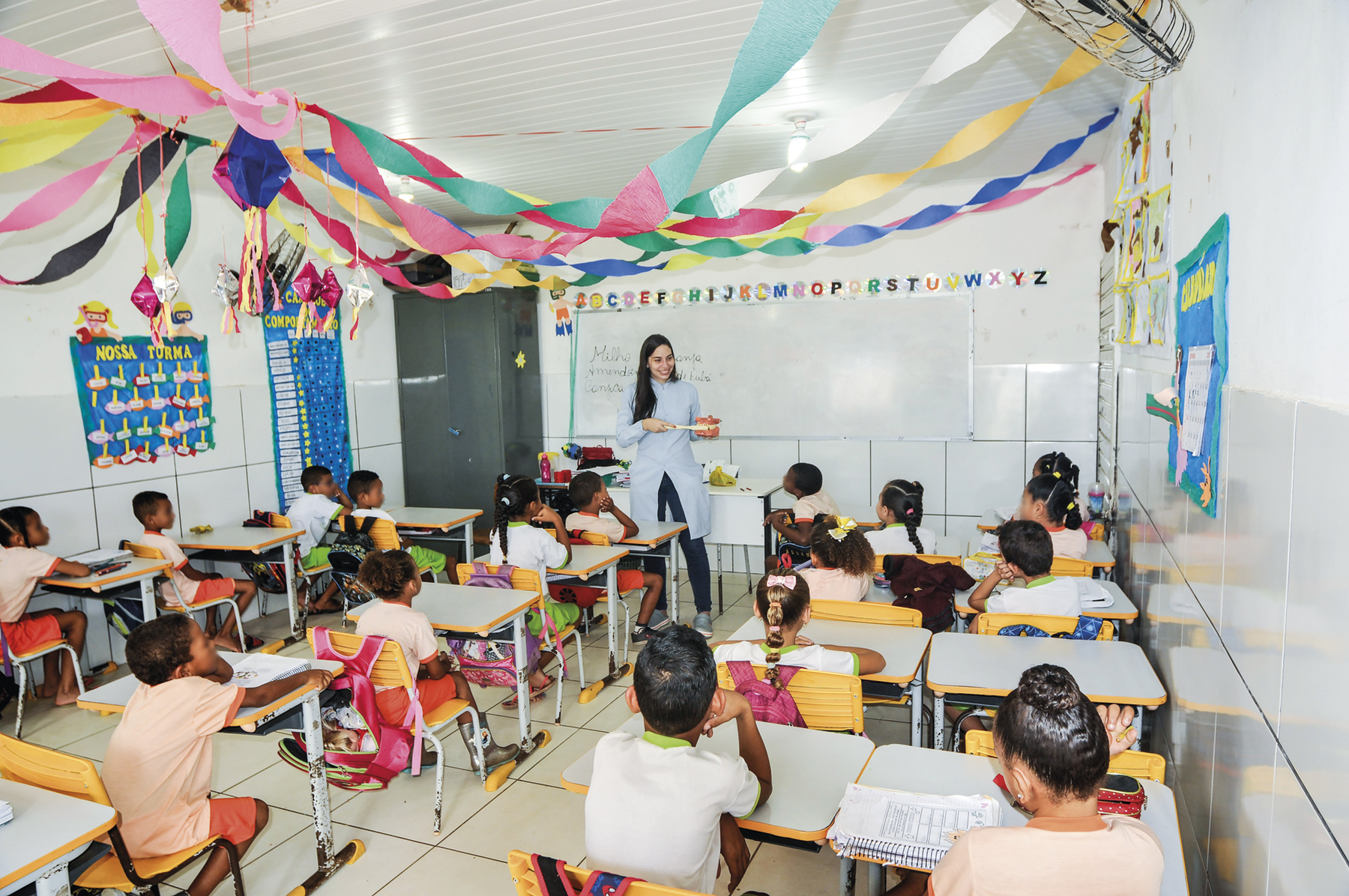 Fotografia. Sala de aula com crianças sentadas em carteiras escolares. Ao fundo, uma professora manuseando um objeto e atrás dela, uma lousa. Há bandeirinhas e um balão pendurados no teto.