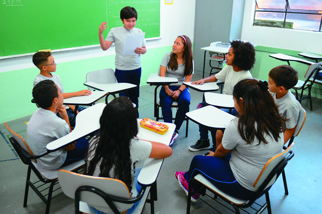 Fotografia. Alunos reunidos em uma sala de aula. Eles estão uniformizados e sentados em carteiras escolares posicionadas em formato circular. Um deles está em pé e lê uma folha de papel.