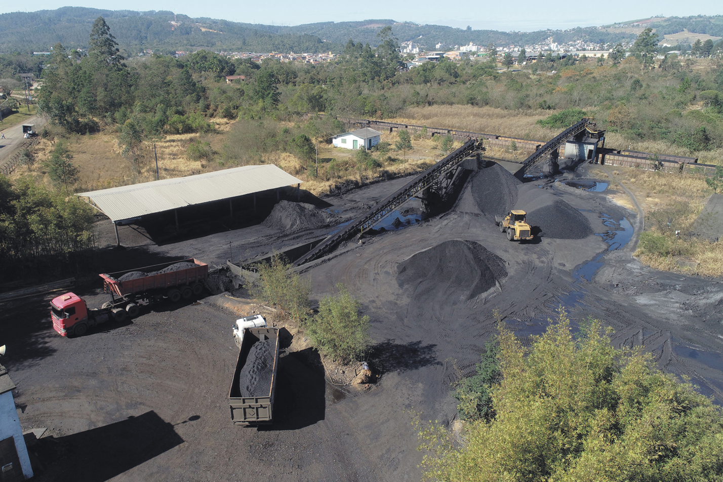 Fotografia. Amontoados de carvão, em uma área com caminhões e gruas. Ao lado há um telhado e ao redor, existem árvores.