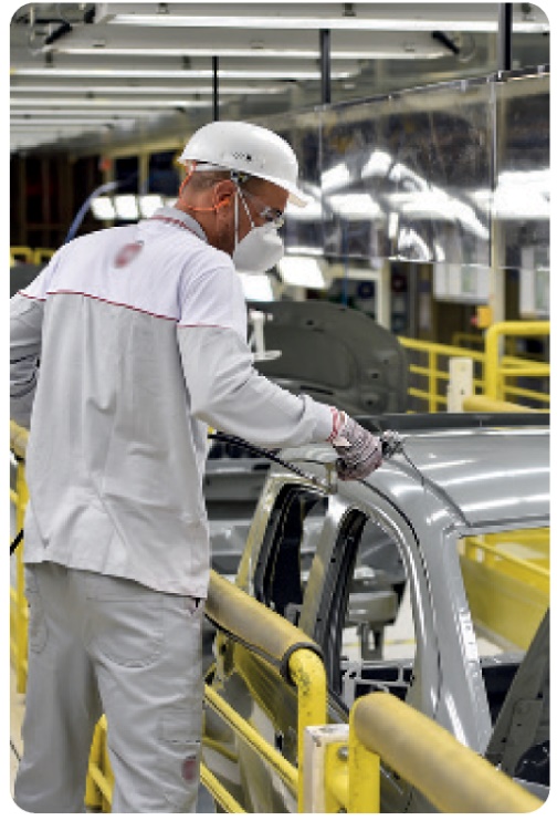 Fotografia. Um homem de uniforme branco com capacete, luvas e máscara, segurando um objeto sobre a carroçaria de um carro em uma linha de produção.