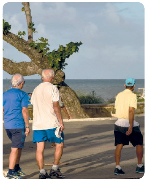 Fotografia. Três idosos caminhando em uma rua à beira-mar. Há uma árvore na calçada.