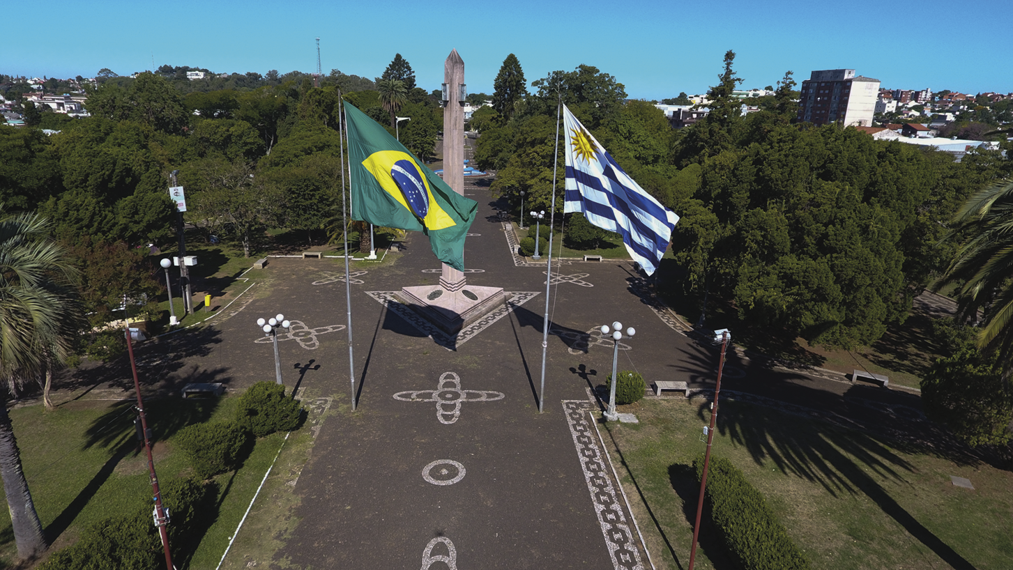 Fotografia. Vista aérea. Um monumento em formato de obelisco no centro de uma praça com a bandeira do Brasil à esquerda e a bandeira do Uruguai à direita. Há árvores ao redor.