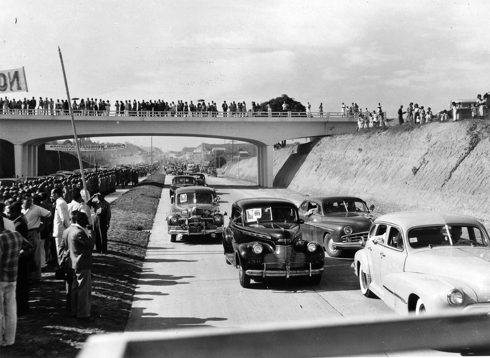 Fotografia em preto e branco. Carros trafegando em uma via com um viaduto ao fundo. Há muitas pessoas na lateral da via e em cima do viaduto observando os carros.