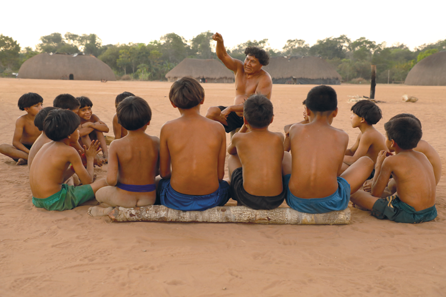 Fotografia. Crianças sentadas em uma área de terra. Há um homem em frente a elas, gesticulando. Ao fundo, moradias indígenas e árvores.