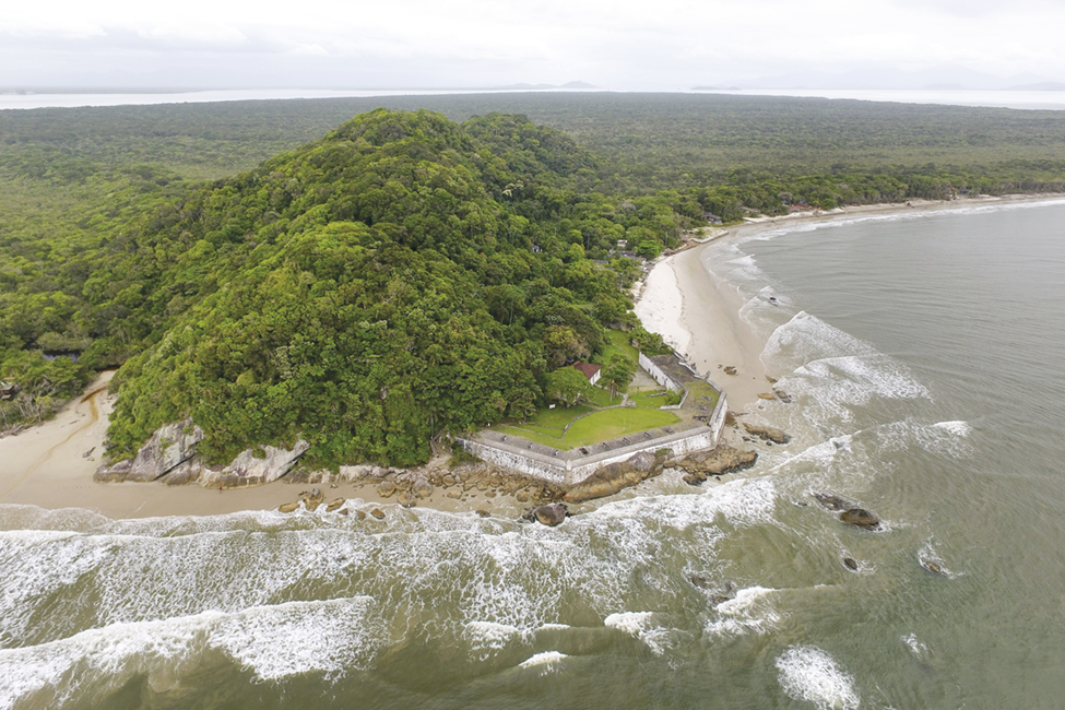 Fotografia. Vista de cima. Uma ilha com vegetação densa e trecho de praia com uma construção e muros à direita.