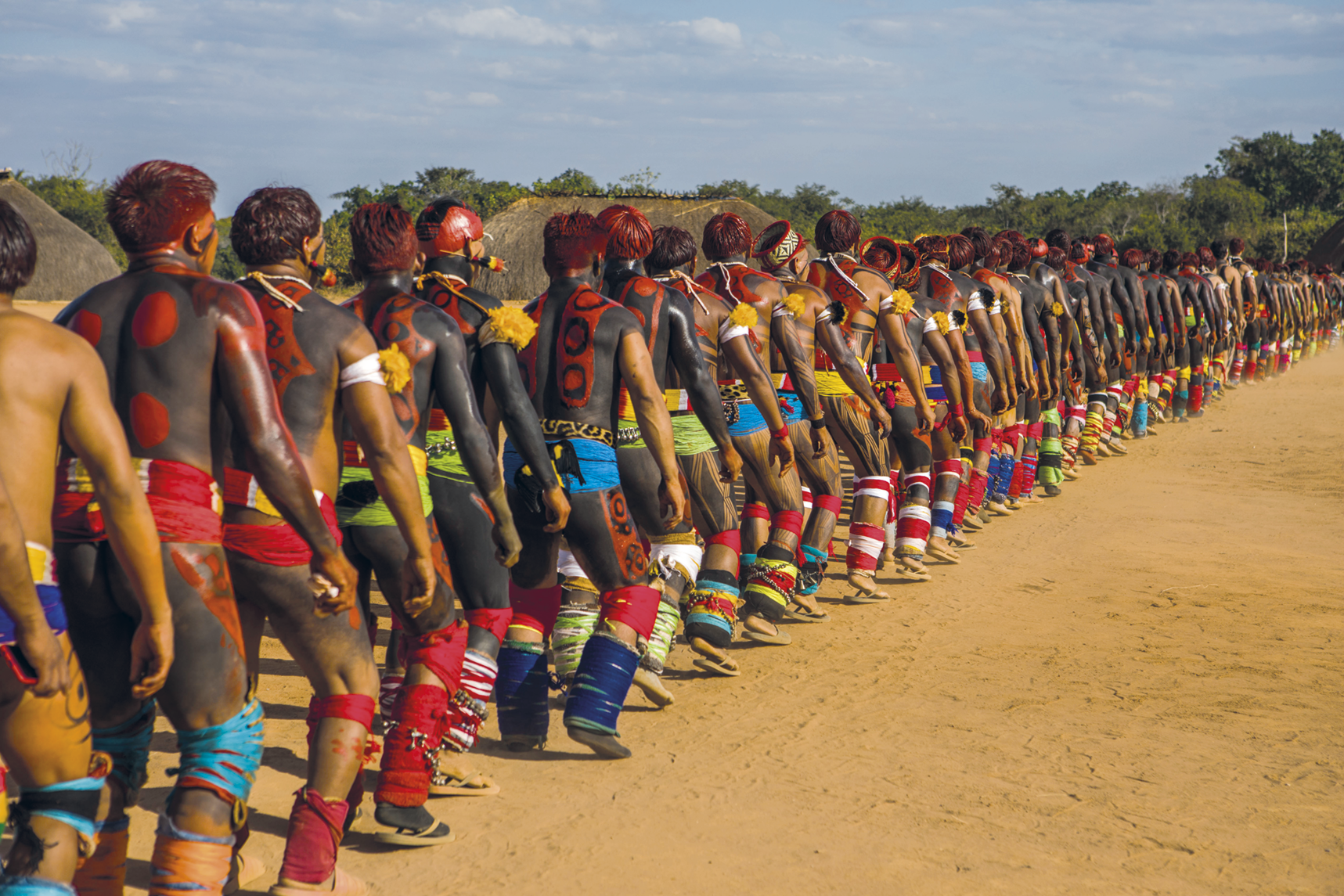 Fotografia. Diversos homens indígenas caminhando enfileirados em uma área de terra. Eles usam pinturas corporais pretas e vermelhas e adornos coloridos ao redor de suas cinturas, joelhos, canelas e braços. Ao fundo há árvores.