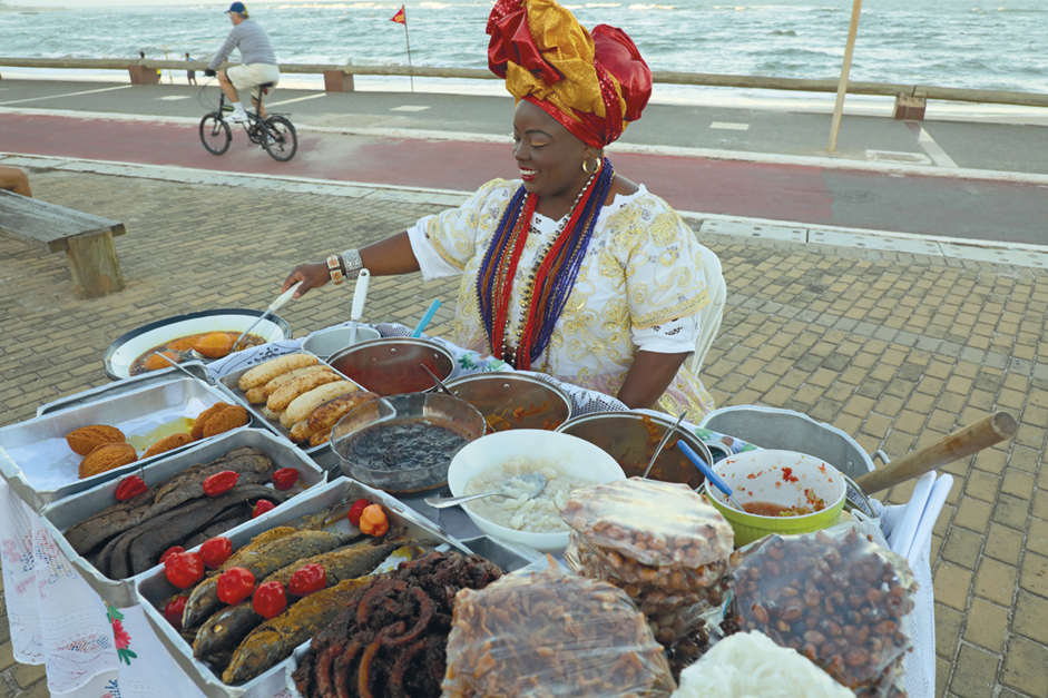 Fotografia. Mulher de vestido e turbante, atrás de uma mesa com travessas com peixes, bolinhos e molhos. Ela está fritando alguns bolinhos. Ao fundo, ciclovia e a praia.