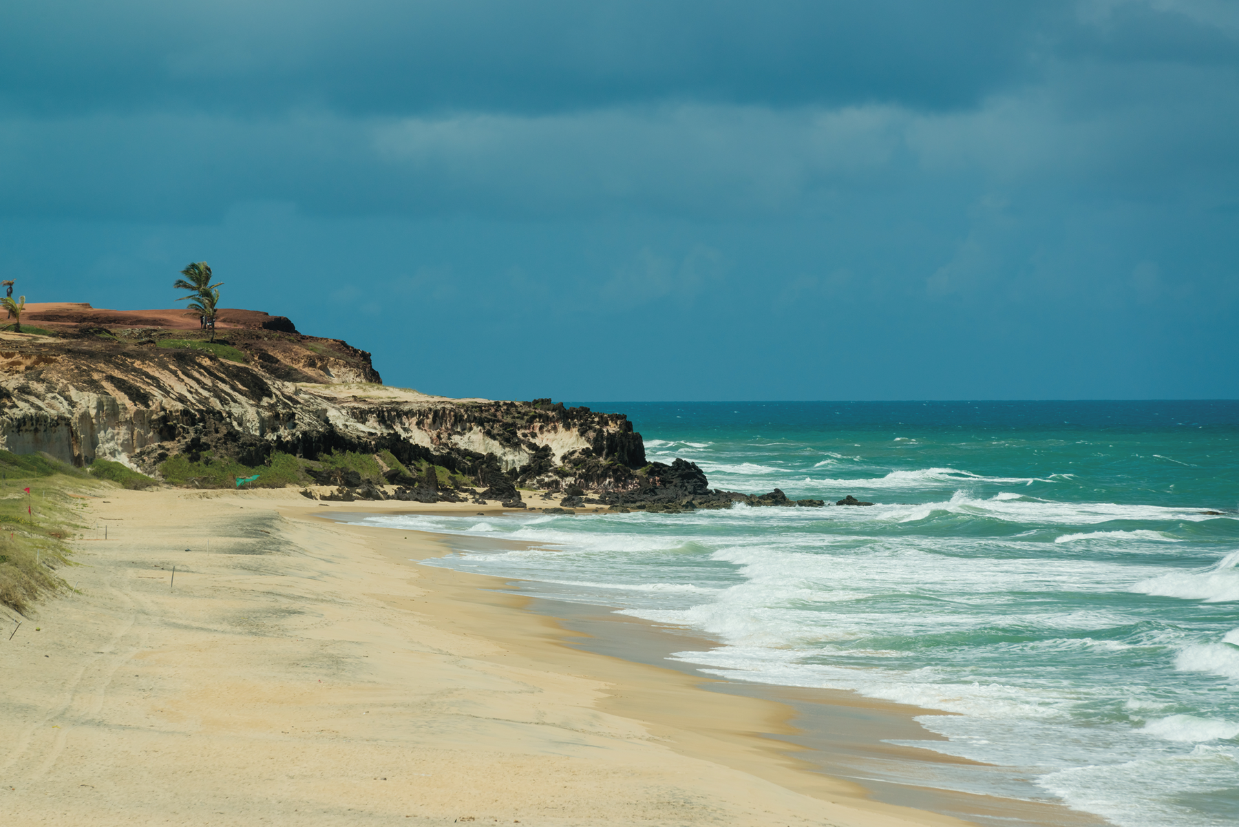 Fotografia. Orla de uma praia com uma pequena elevação rochosa na faixa de areia, onde há algumas palmeiras. No mar, ondas.