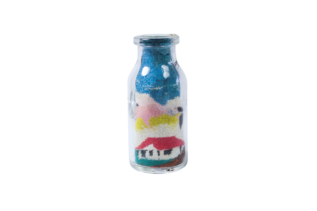 Fotografia. Uma garrafa de vidro com areia colorida compondo a figura de uma casa, árvores e céu azul.