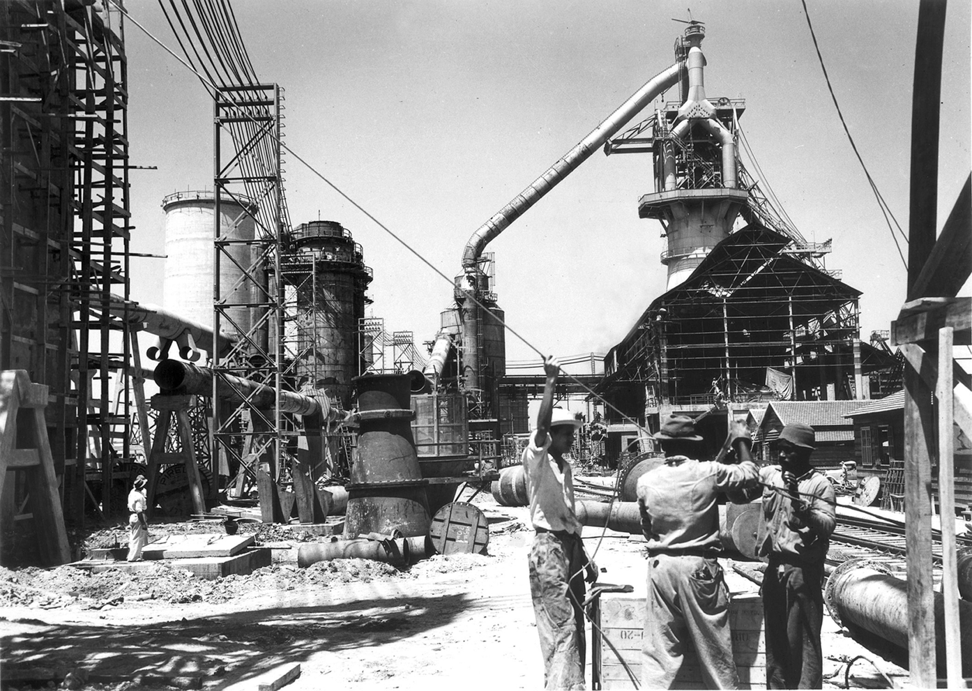 Fotografia em preto e branco. Alguns homens reunidos em uma construção. Há colunas e estruturas de metal interligadas e um galpão ao fundo, à direita.