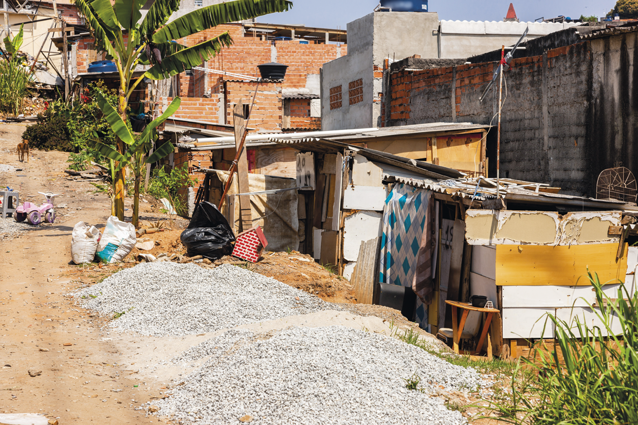 Fotografia. Moradias improvisadas de tábuas em uma rua sem pavimentação. Há pedras em frente a elas e ao fundo, casas precárias e bananeiras.