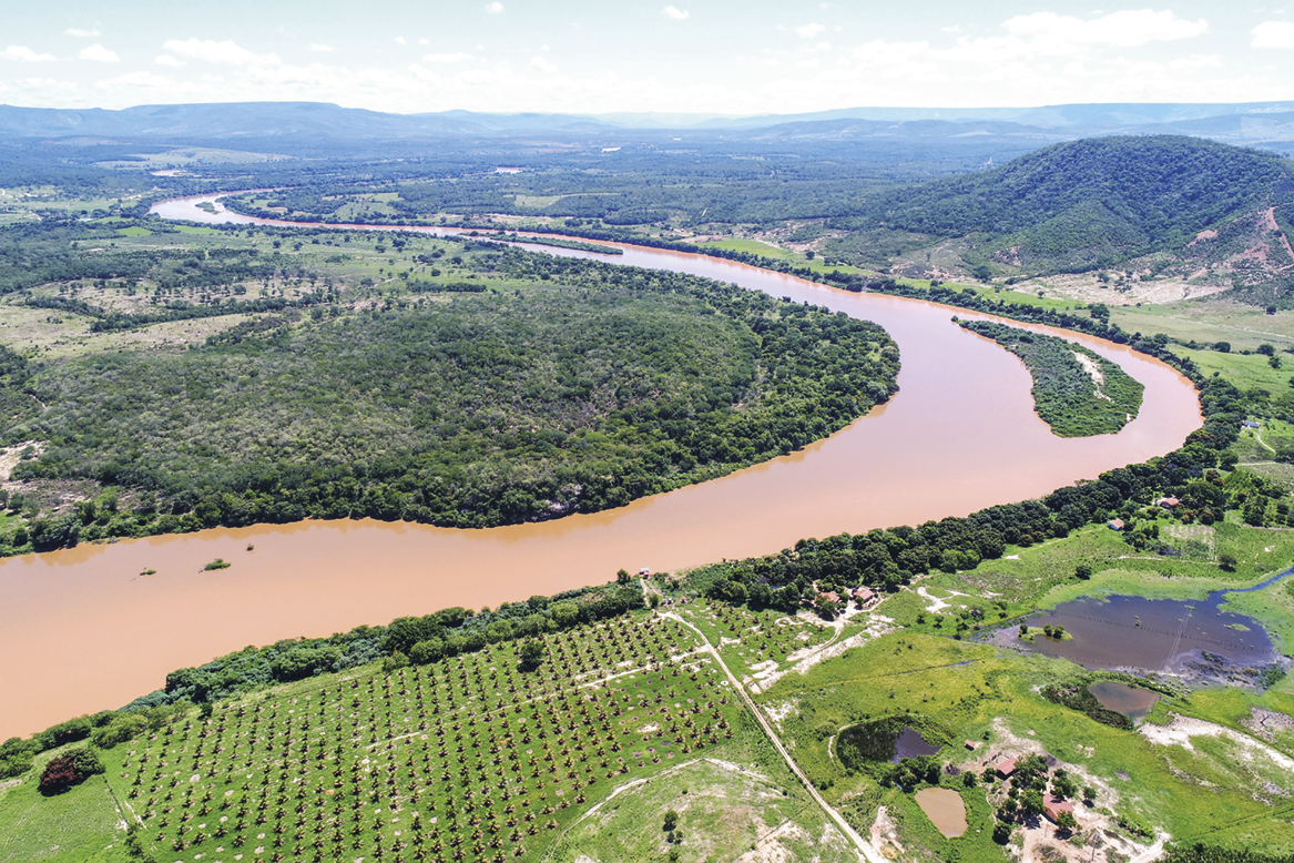 Fotografia. Vista de cima. Curso sinuoso de um rio com as margens com vegetação. Abaixo, área de agricultura.