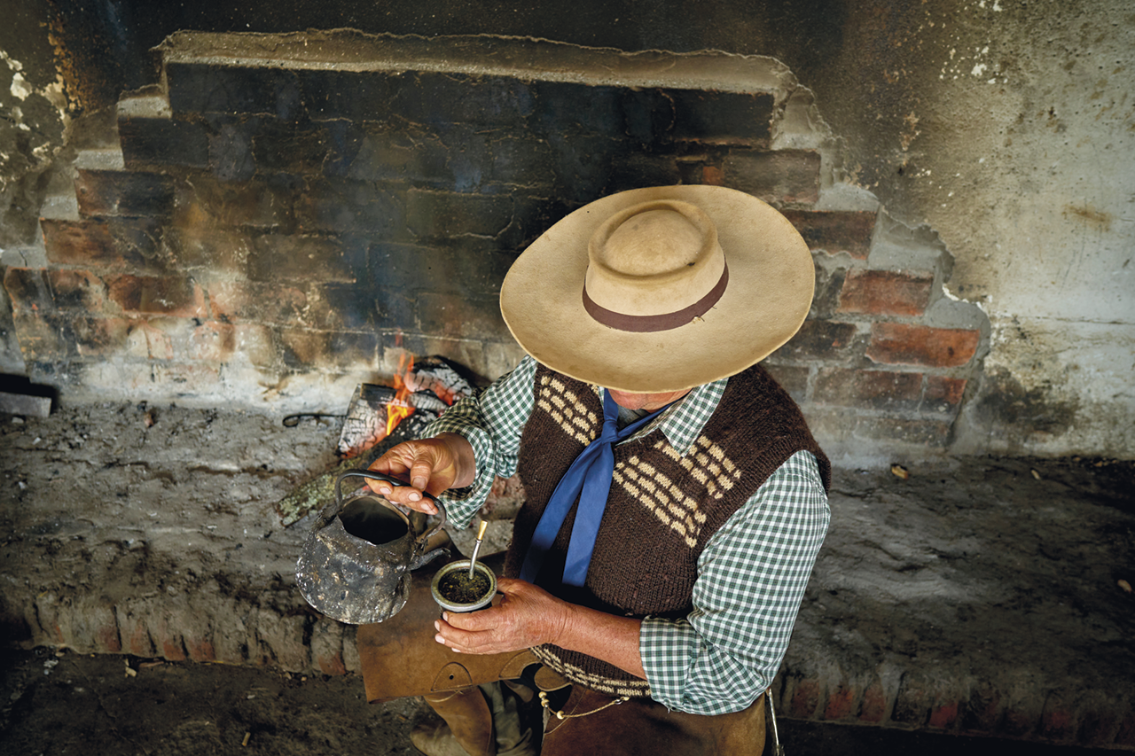 Fotografia. Vista de cima. Homem de chapéu, lenço, colete, e camisa xadrez servindo água em uma cuia de chimarrão. Ao fundo, parede e uma fogueira.