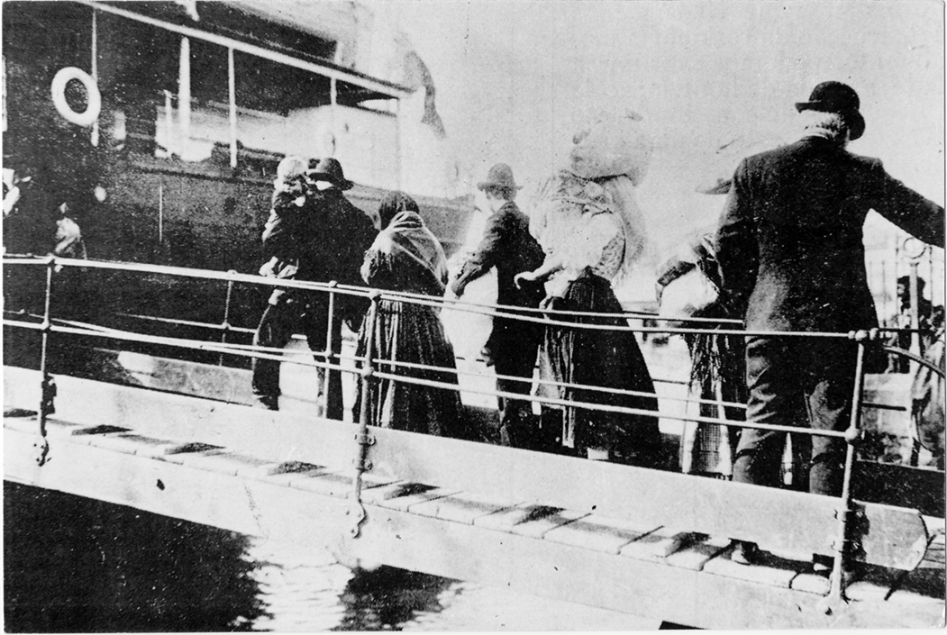 Fotografia em preto e branco. Homens e mulheres subindo uma rampa em direção a um navio. Eles carregam crianças e trouxas.