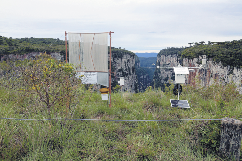 Fotografia. Uma antena e um equipamento retangular no alto de um morro coberto por vegetação. Ao fundo, morros de topo plano formando uma grande fenda entre eles.