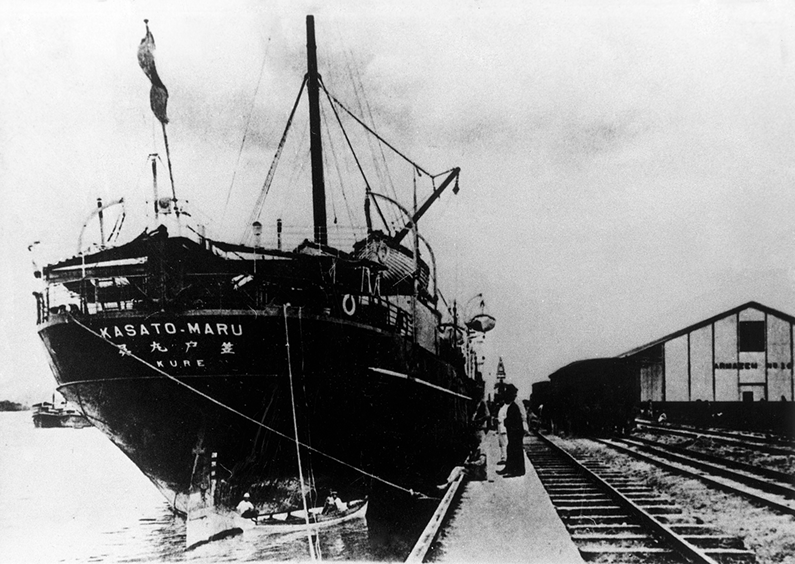 Fotografia. Um navio ancorado ao lado de trilhos de trem e de uma construção.