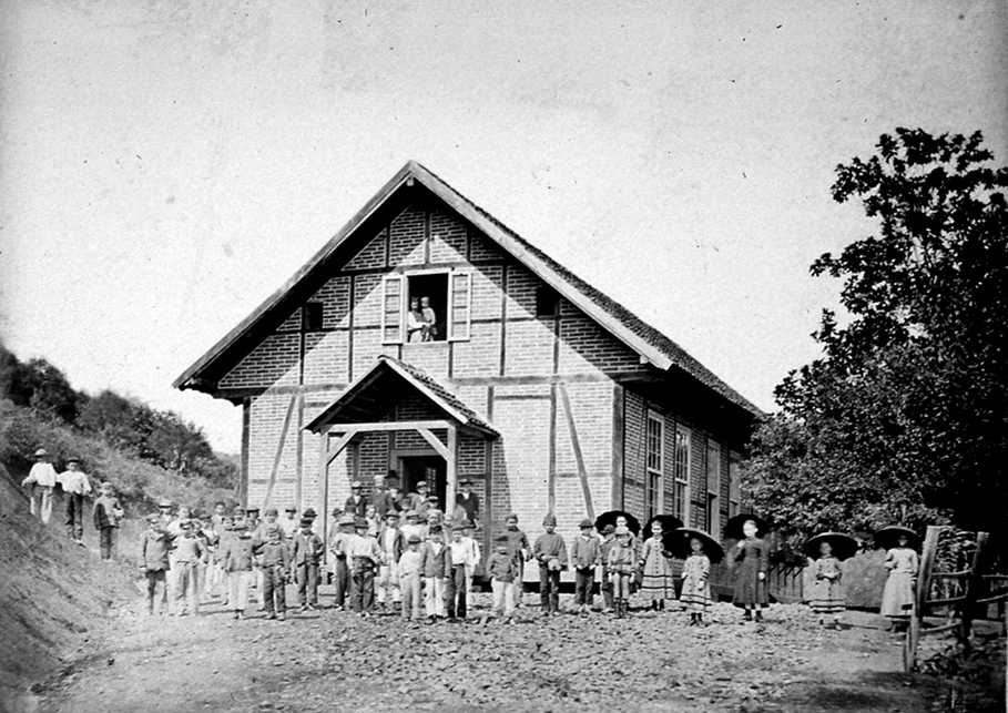 Fotografia em preto e branco. Muitas pessoas reunidas em frente a uma construção de madeira com árvores ao redor.