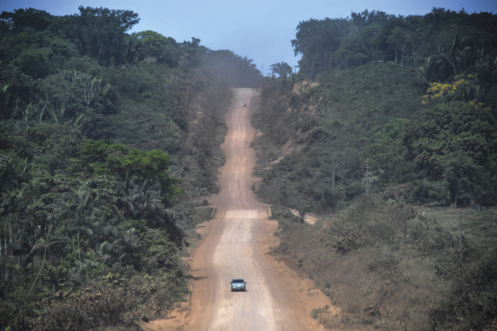 Fotografia. Uma estrada de terra cercada por uma floresta. Há um carro trafegando.