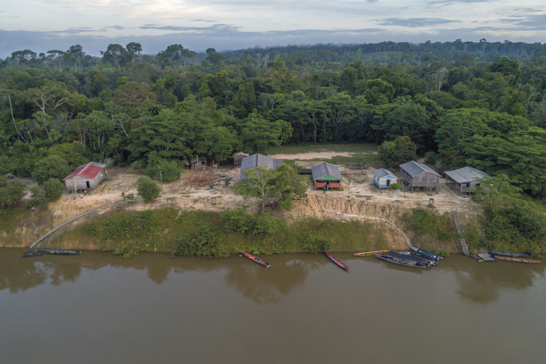 Fotografia. Vista de cima. Uma comunidade na margem de um rio. Há casas espalhadas em áreas desmatadas e ao fundo, floresta.