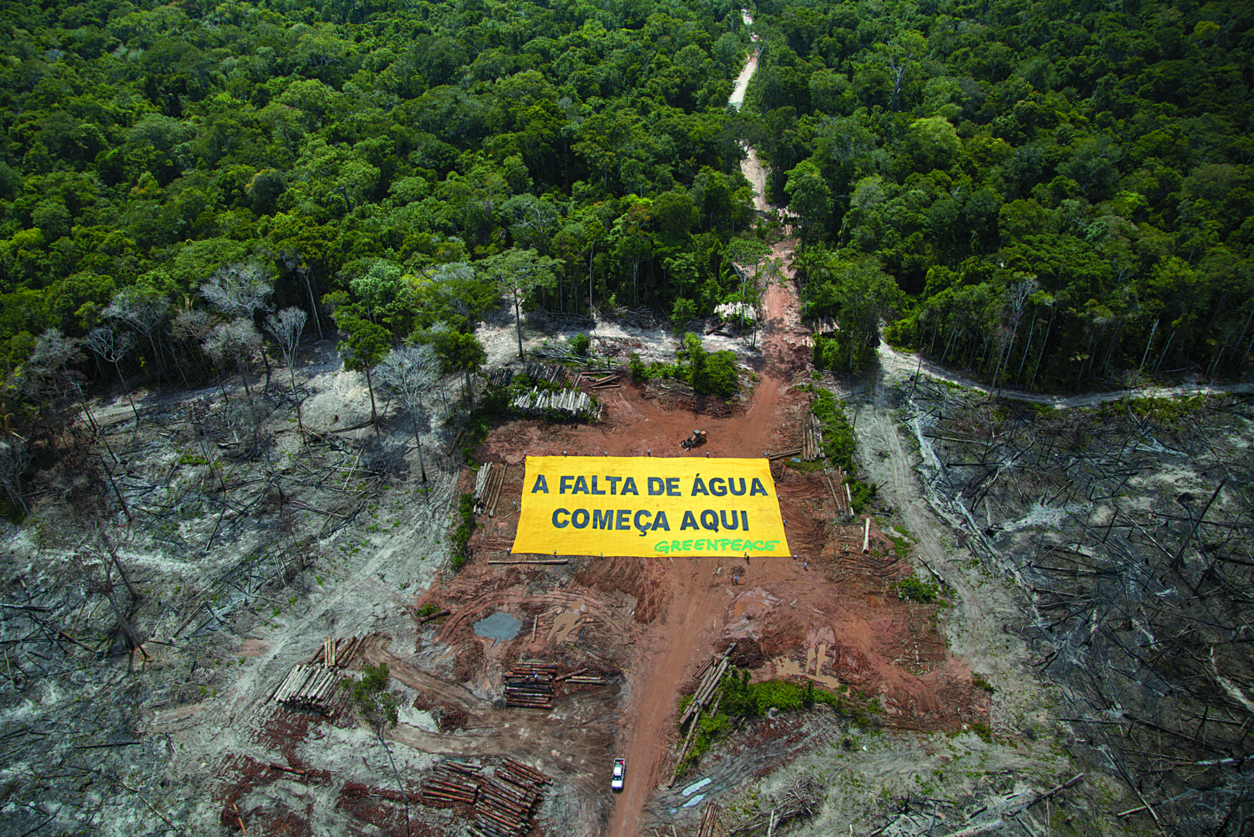 Fotografia. Vista aérea. Área desmatada, de terra com uma faixa com a inscrição: A FALTA DE ÁGUA COMEÇA AQUI. Ao lado há áreas queimadas e ao fundo, uma floresta.