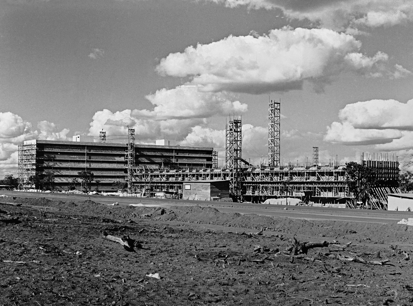 Fotografia em preto e branco. Prédios com andaimes e estruturas de ferro em frente a eles. No primeiro plano há um campo aberto.