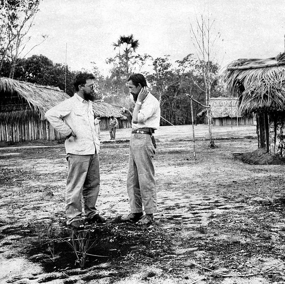 Fotografia em preto e branco. Dois homens conversando frente a frente em uma área aberta. Ao redor há construções de palha.