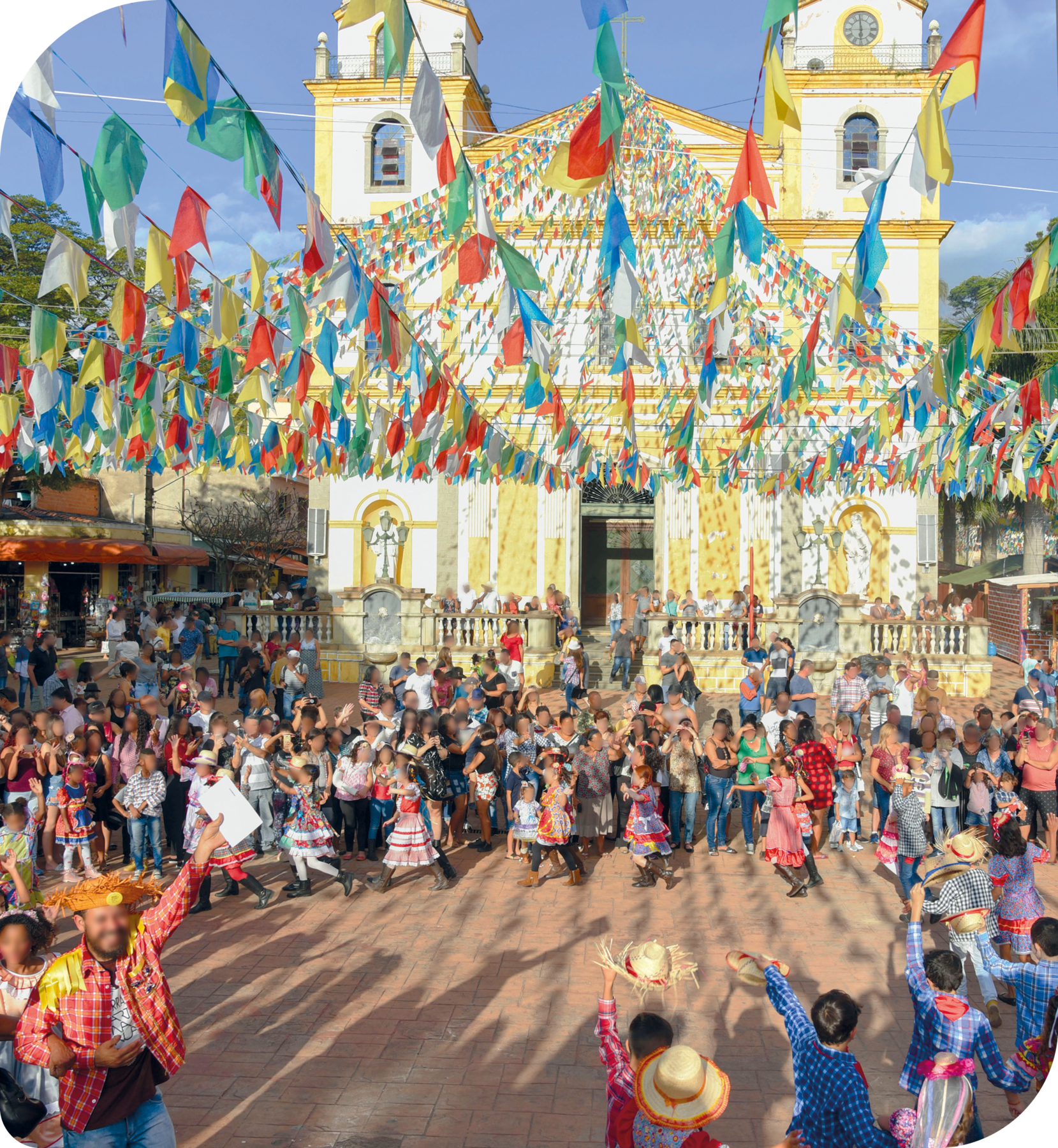 Fotografia. Muitas pessoas reunidas em círculo em frente a uma igreja. Há pessoas dançando quadrilha e outras que assistem. Na parte superior, bandeirinha coloridas penduradas. A igreja ao fundo é branca com detalhes amarelos.