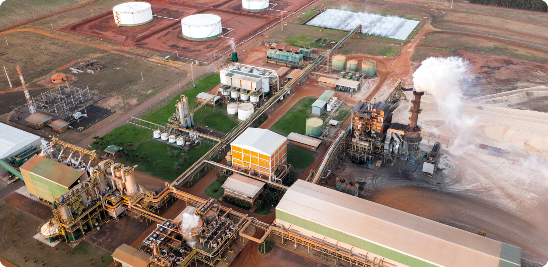 Fotografia. Vista de cima. Uma usina com galpões, silos e estruturas de ferro interligadas. Há uma torre expelindo fumaça.