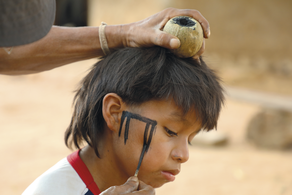 Fotografia. Destaque para uma pessoa fazendo pintura no rosto de uma criança indígena. Ela passa o pincel na bochecha do menino e segura um potinho com tinta acima da cabeça dele.