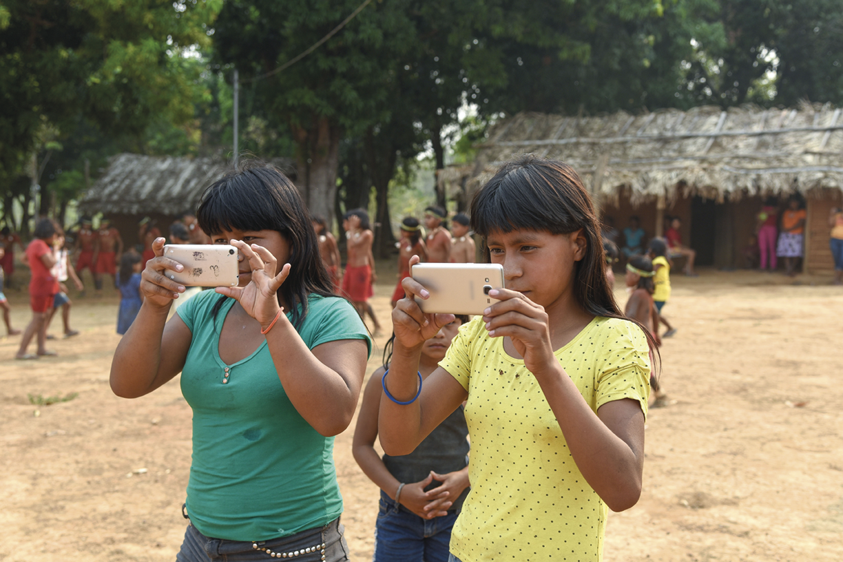 Fotografia. Duas mulheres indígenas com celulares apontados para frente. Atrás delas, há outros indígenas e moradias de palha. Ao fundo há árvores.