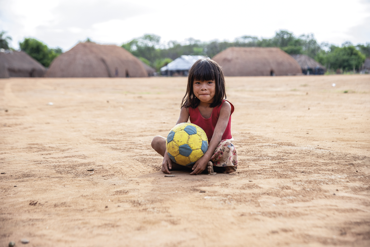 Fotografia. Criança de cabelos médios sentada em um campo de terra. Ela segura uma bola de futebol em seu colo. Ao fundo há moradia indígenas.
