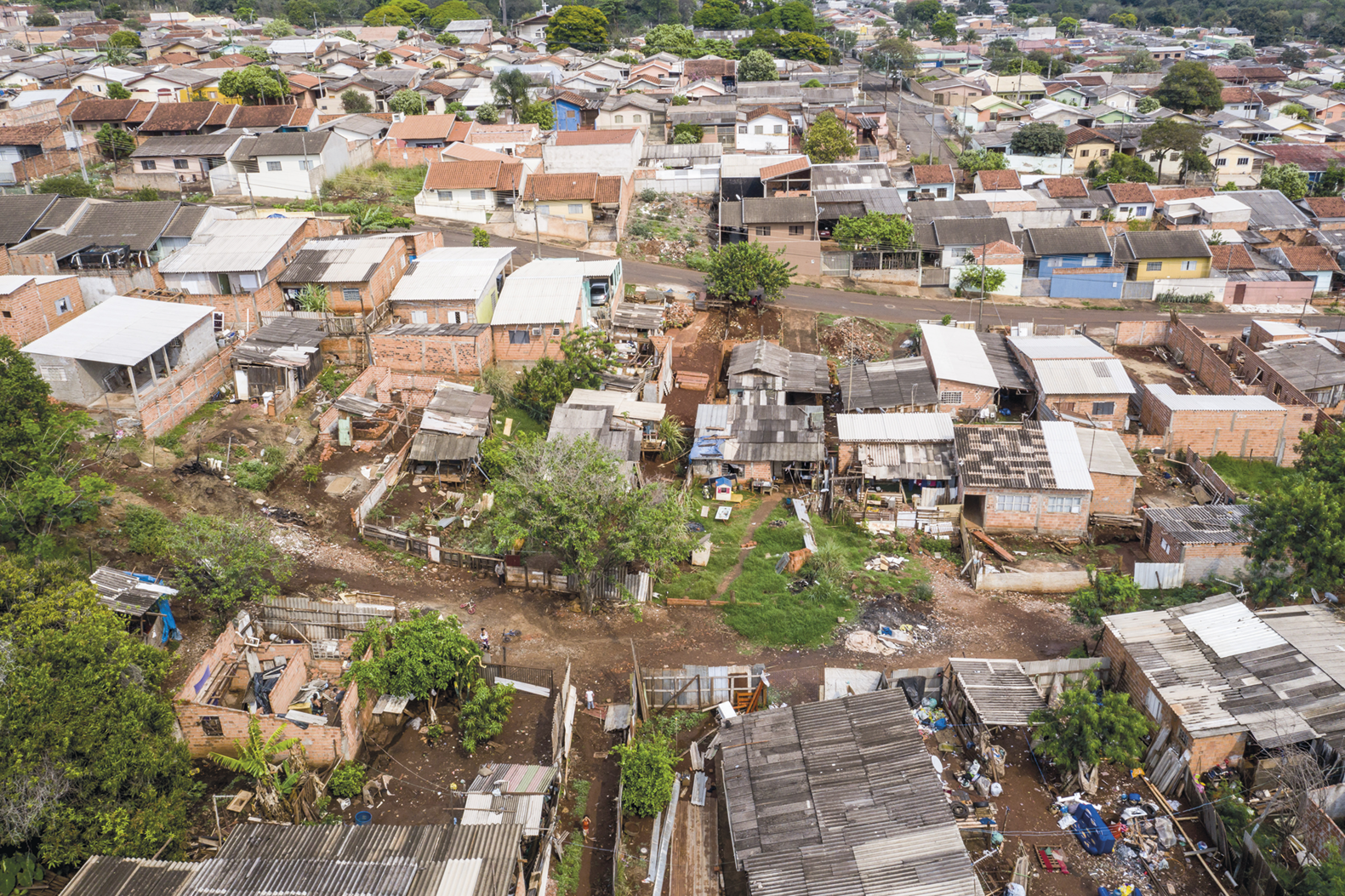 Fotografia. Vista aérea. Diversas casas amontoadas em uma área de terra com árvores. Há um terreno com lixo.