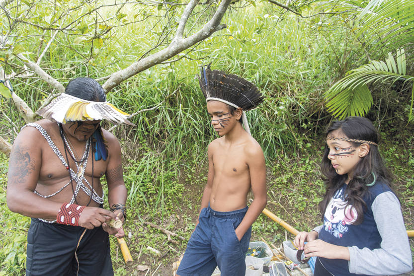 Fotografia C. À esquerda, homem de cocar com colares e pulseiras, manuseando um objeto. Em frente a ele, há uma menina e um menino também de cocar. Há uma área com vegetação atrás deles.