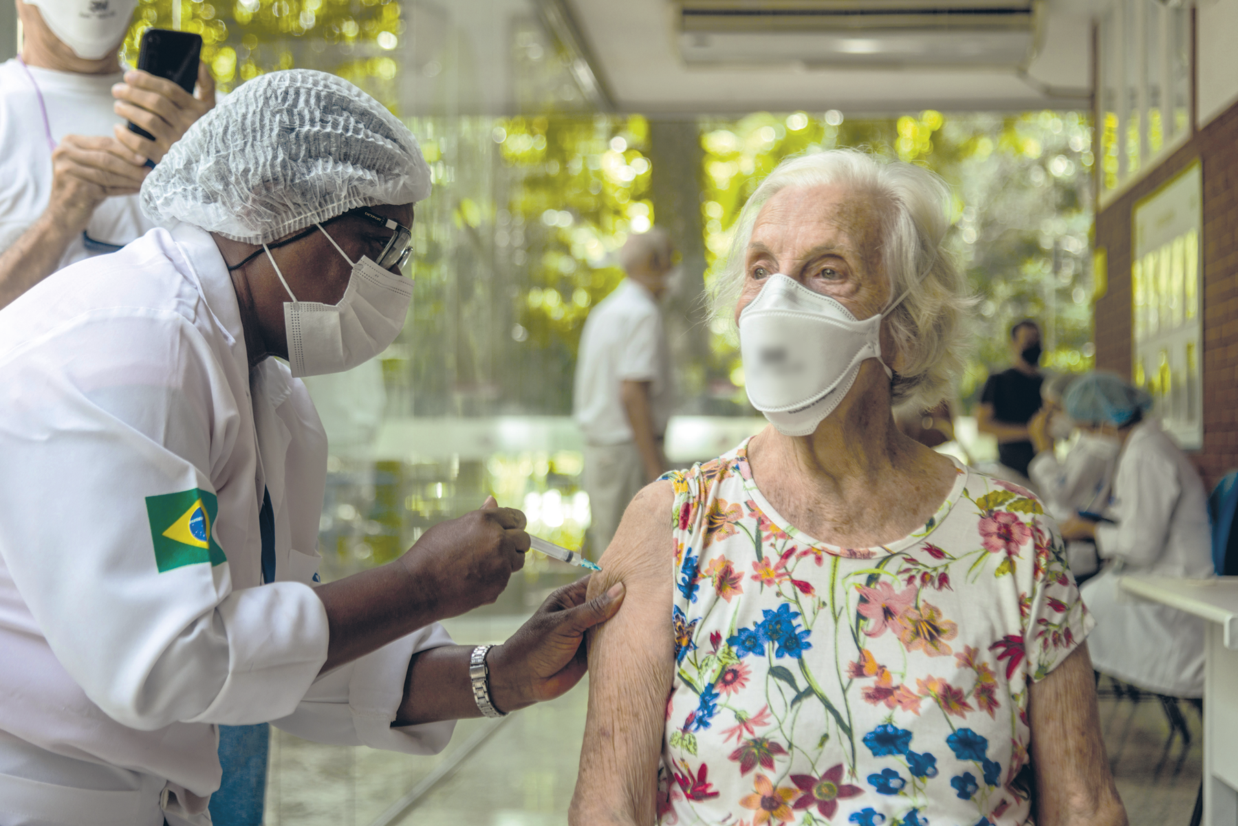 Fotografia. Destaque para uma idosa sendo vacinada. A mulher que aplica a vacina em seu braço usa touca, máscara e avental. A senhora também usa máscara. Ao fundo há mais pessoas.