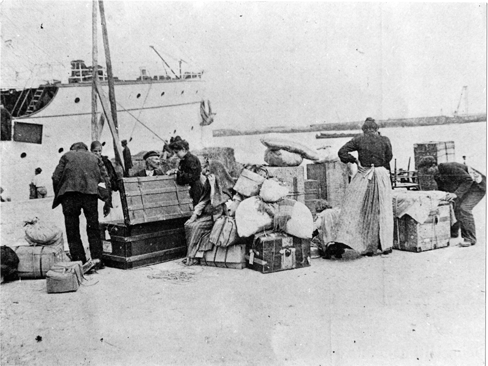 Fotografia em preto e branco. Algumas pessoas entre malas e caixas. Ao fundo há um navio.