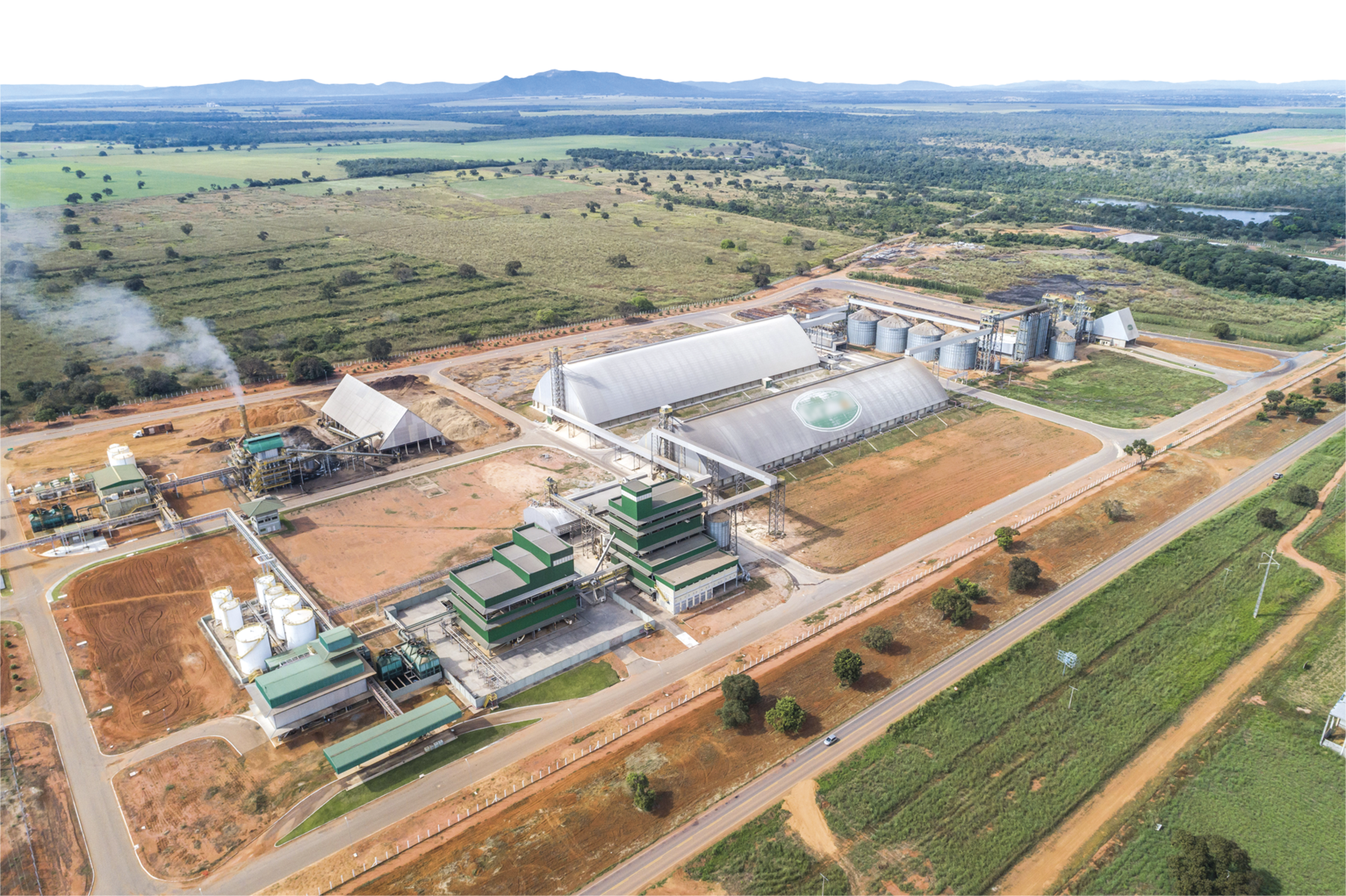 Fotografia. Vista de cima. Área com galpões prédios, silos e estruturas de metal entre áreas de terra. Há uma torre expelindo fumaça e ao redor, áreas cobertas por vegetação.