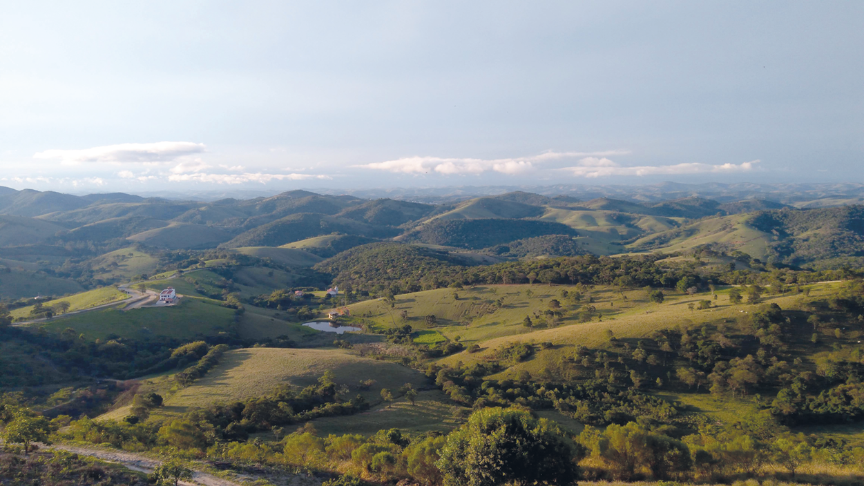 Fotografia. Vista de cima. Morros e cobertos por vegetação rasteira e com algumas árvores.