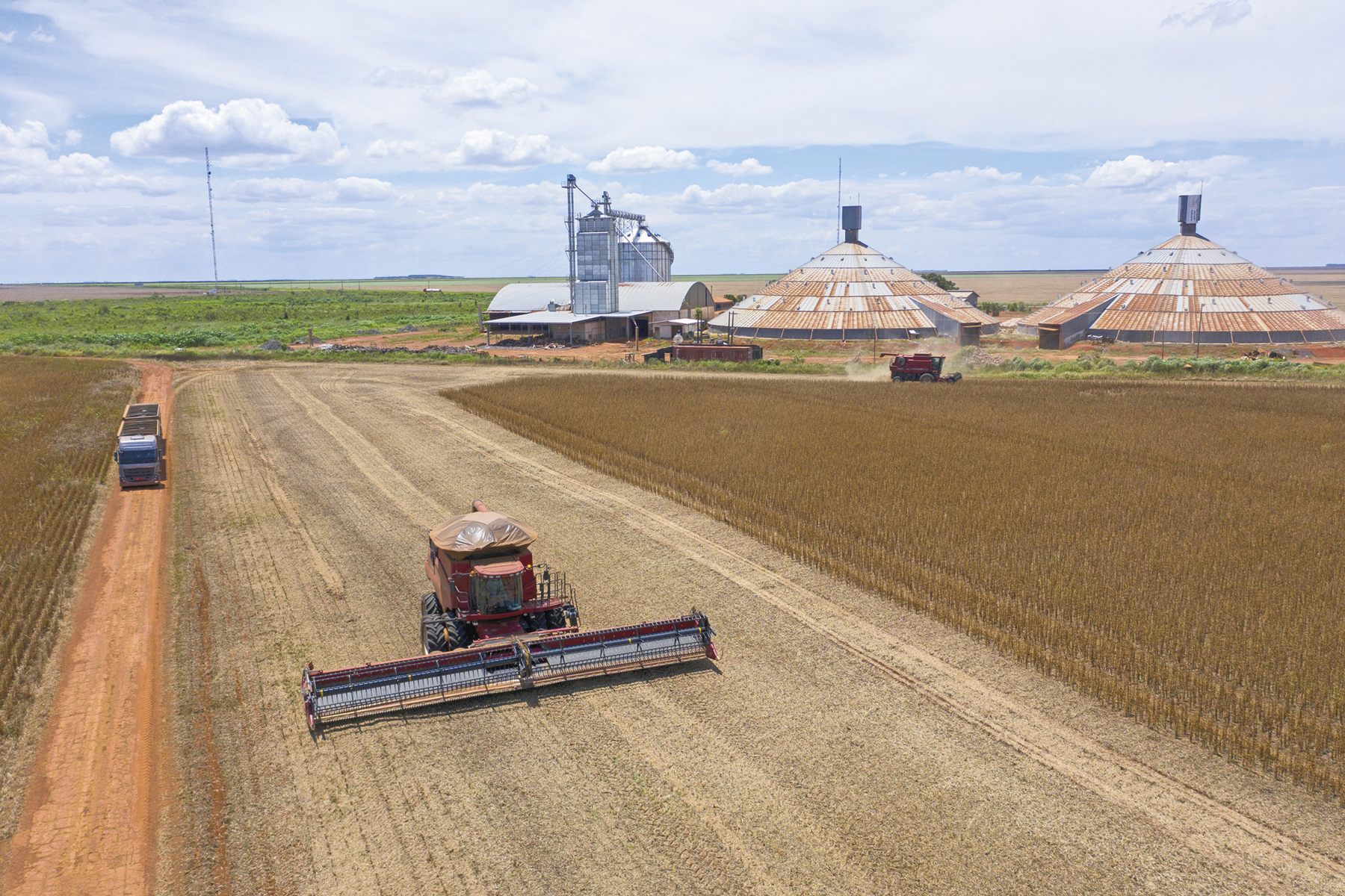 Fotografia. Uma colheitadeira passando por uma extensa plantação de soja. Ao fundo, silos e galpões de metal.