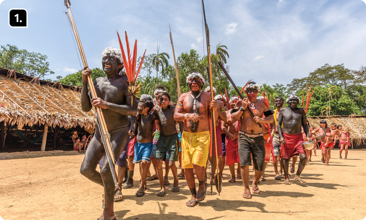 Fotografia 1. Indígenas usando adornos com penas nas cabeças, empunhando lanças. Eles estão caminhando para frente. Ao redor há moradias e árvores.