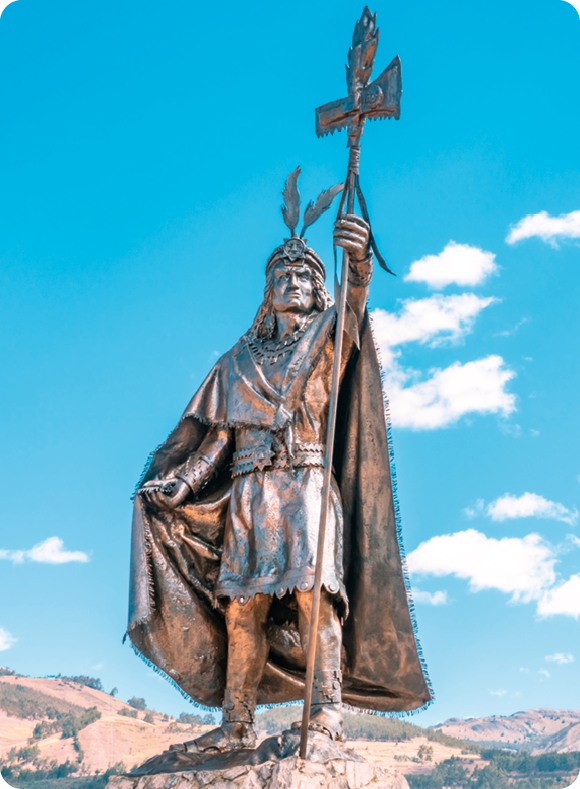 Fotografia. Monumento de um homem de túnica e capa com um adorno na cabeça empunhando um estandarte com um formato de uma cruz.