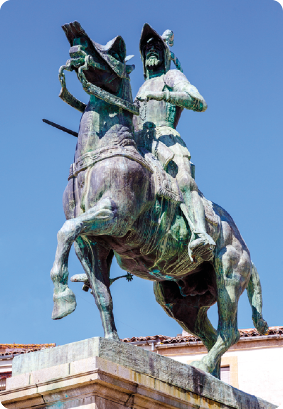 Fotografia. Monumento com um homem de chapéu montado em um cavalo que usa um adorno na cabeça.