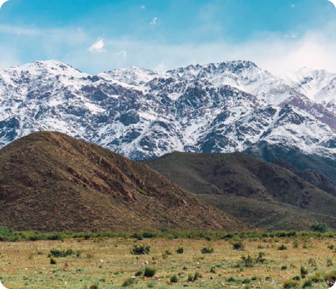 Fotografia. Conjunto de montanhas com neve, em frente morros menores e área plana coberta por vegetação.