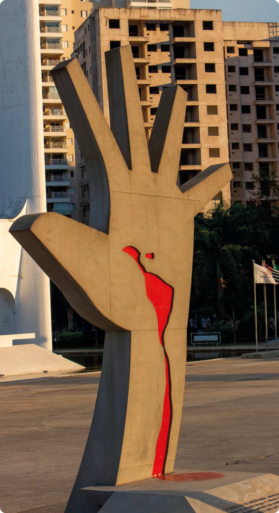 Fotografia. Escultura de uma mão espalmada com a silhueta da América Latina em vermelho, com um traçado que estende até o pulso. Ao fundo, prédios.