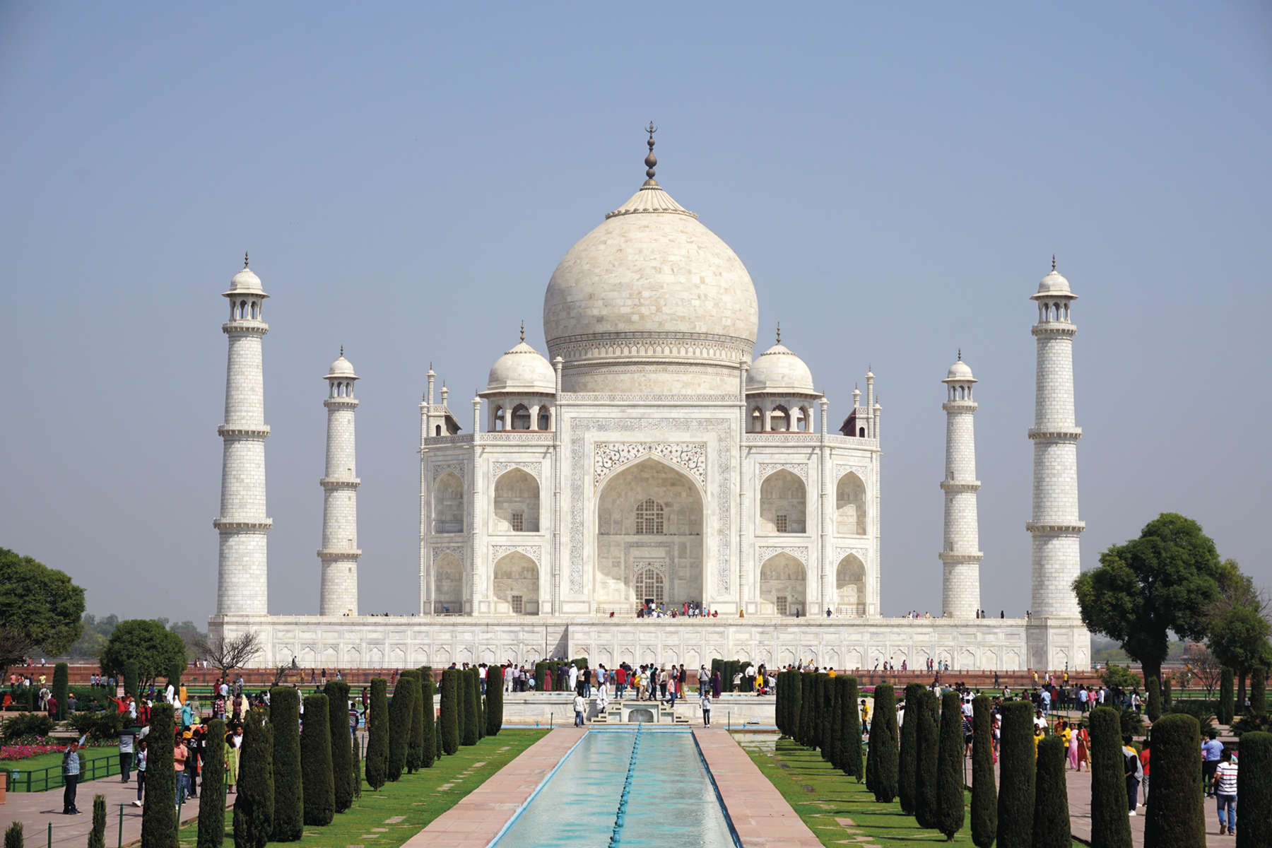 Fotografia. Taj Mahal, construção ampla com uma grande cúpula no topo ao centro e torres finas ao redor. Em frente a uma piscina retangular com fileiras de árvores e pessoas entre elas.