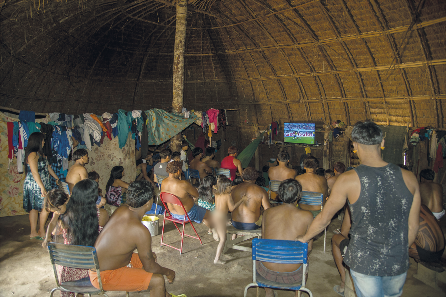 Um grupo de indígenas reunidos em uma moradia indígena, assistindo uma partida de futebol em uma televisão que está ao fundo. A maioria deles está sentada em cadeiras, outros estão em pé. Ao fundo há roupas penduradas.