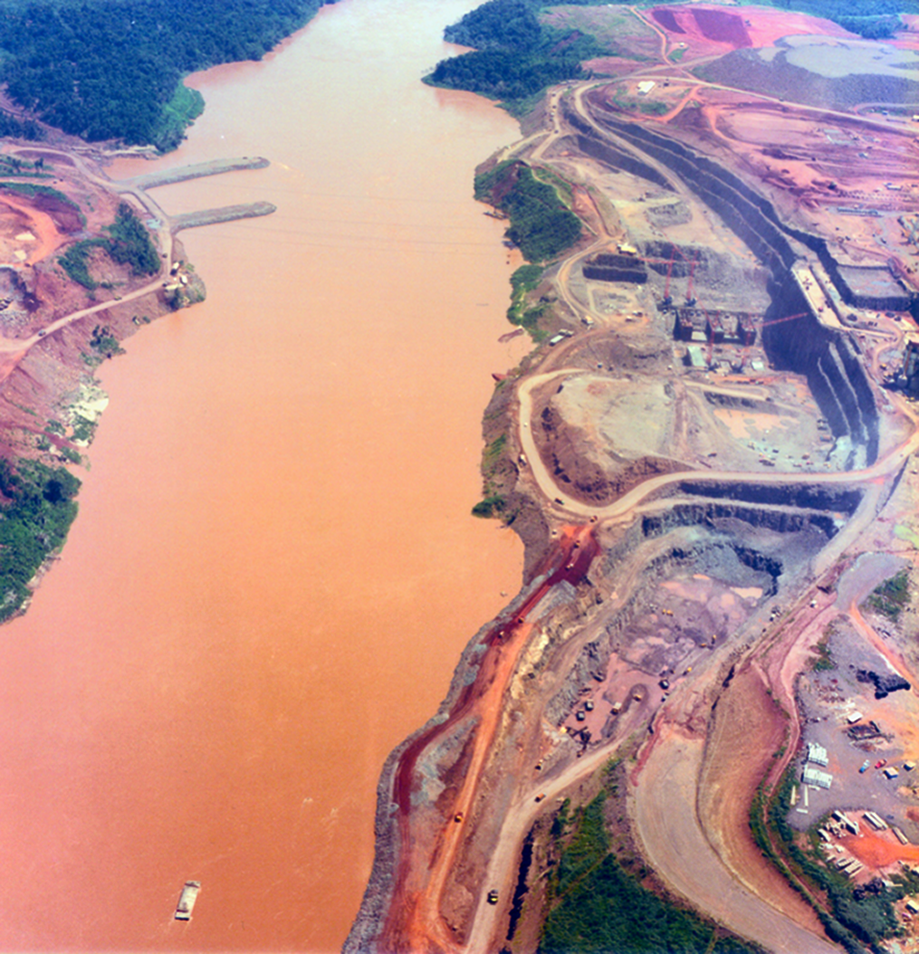 Fotografia. Vista aérea. Curso de um rio marrom entre área desmatada com construções nas margens.
