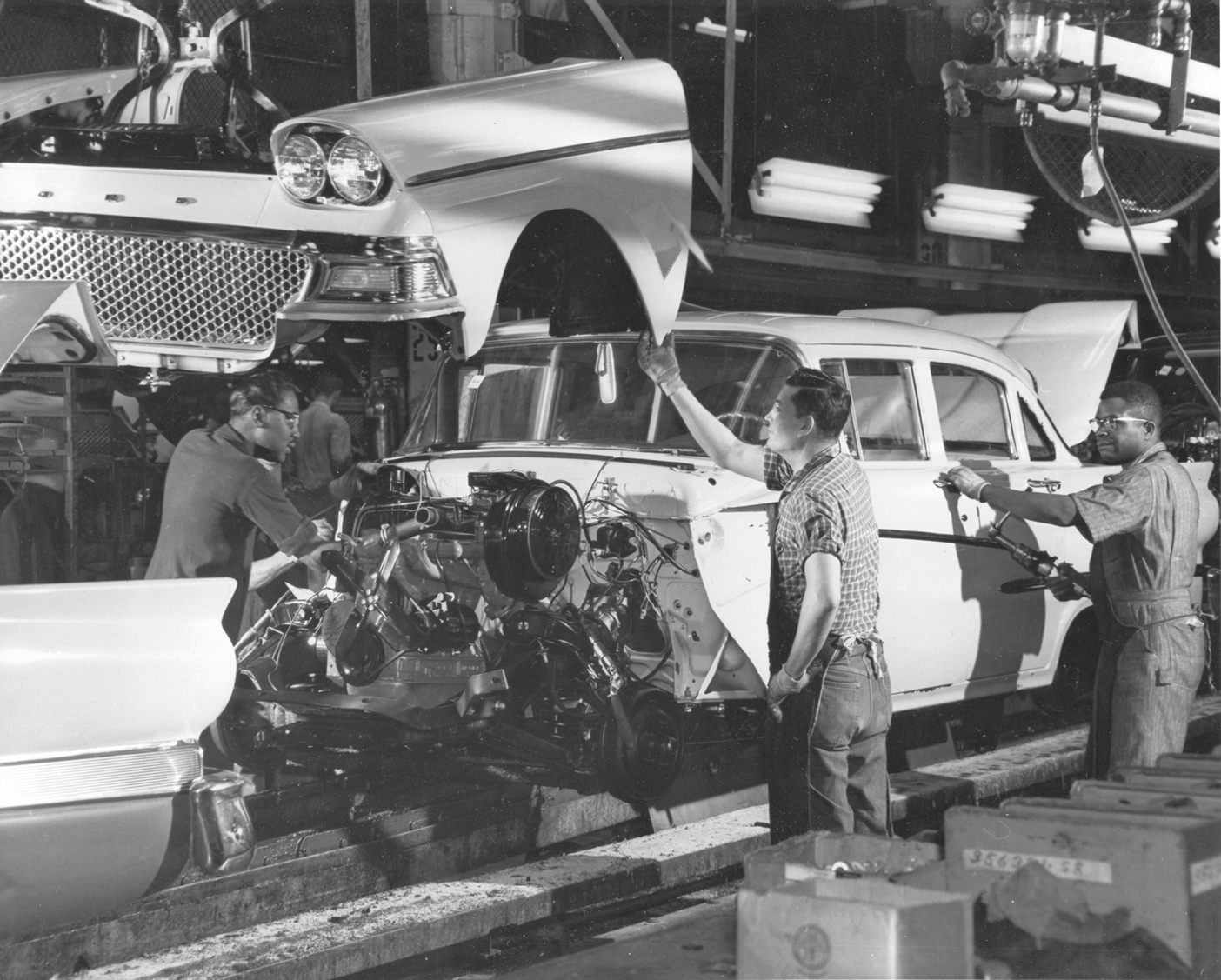 Fotografia em preto e branco. Homens trabalhando em uma linha de montagem, posicionando a carroçaria de um automóvel sobre o motor. Há estruturas e equipamentos ao lado deles.