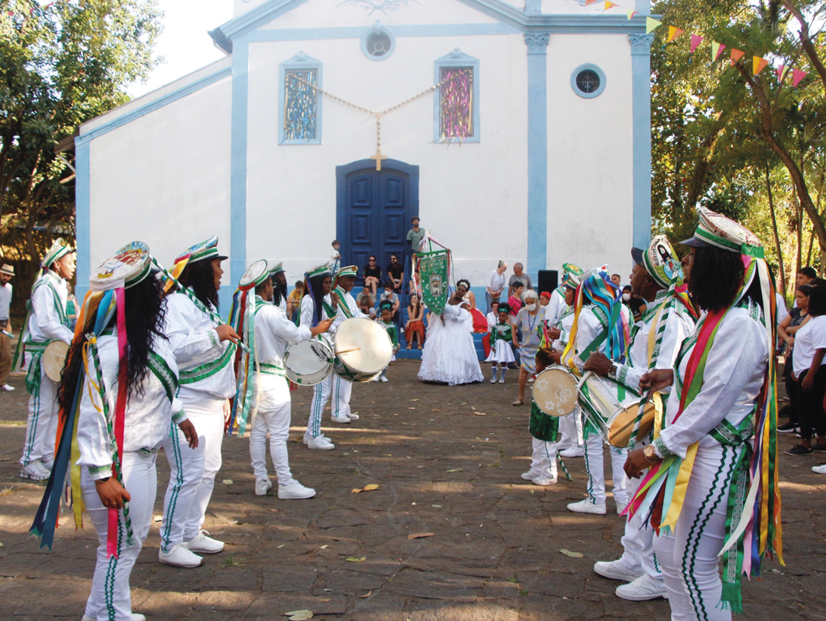 Fotografia. Pessoas com uniformes brancos com fitas coloridas e chapéus tocando instrumentos e formando um corredor em frente a uma igreja, que está ao fundo. Em frente à igreja uma pessoa de vestido branco desfila segurando um estandarte verde. Há mais pessoas ao redor dela.