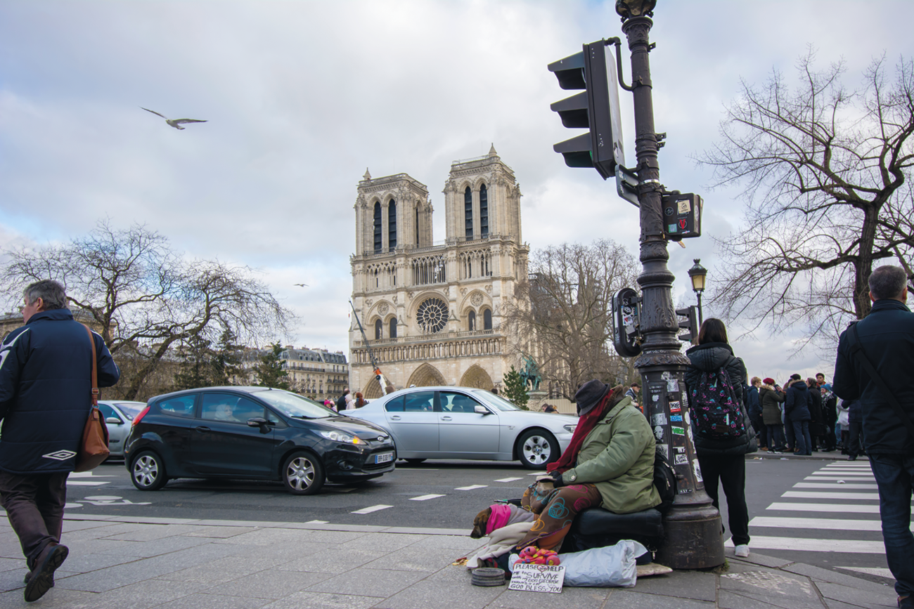 Fotografia. Uma pessoa sentada ao lado de um semáforo. Há roupas e bolsas ao redor dela. Ao lado, carros trafegando, pessoas na calçada, e ao fundo, uma igreja com duas torres e árvores secas.