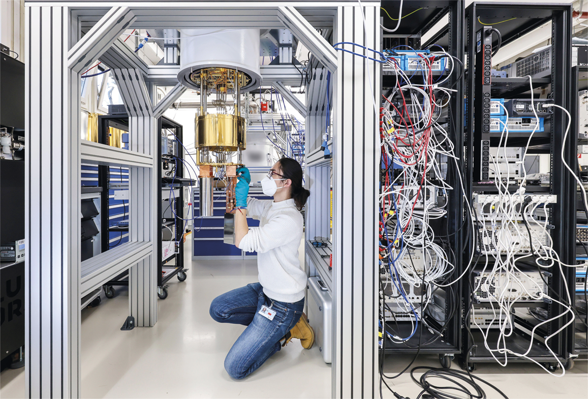 Fotografia. Uma mulher de luvas e máscara manuseando cabos em um equipamento que está suspenso em uma sala com estruturas de metal. À direita há diversos cabos.